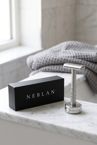 Coffret noir Neblan couché et rasoir de sûreté Neblan 110 sur son support, au bord d'un lavabo en marbre blanc